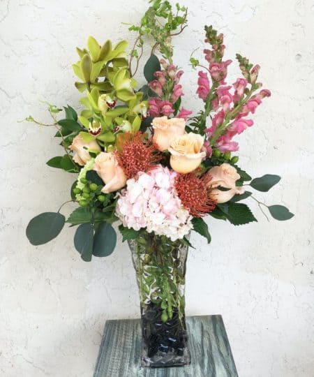Island Cabana features crisp green cymbidiums, pink snapdragons, orange protea, peach roses, fluffy pink hydrangea, green hypericum, and curly willow accented with silver dollar eucalyptus and river rock in this tall glass marbled-effect vase. 