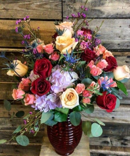 Red and peach roses, lavender hydrangea, blue thistle and more pack into this gorgeous keepsake red vase
