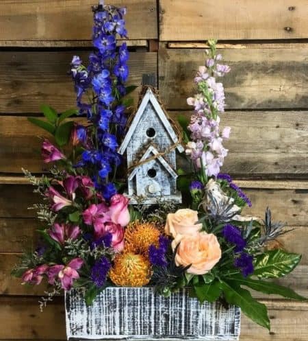  beautiful mix of bright colored flowers, including blooming alstromeria, bright blue delphinium, soft roses, statice and assorted greenery. Perched with a rustic bird making itself a new home. 
